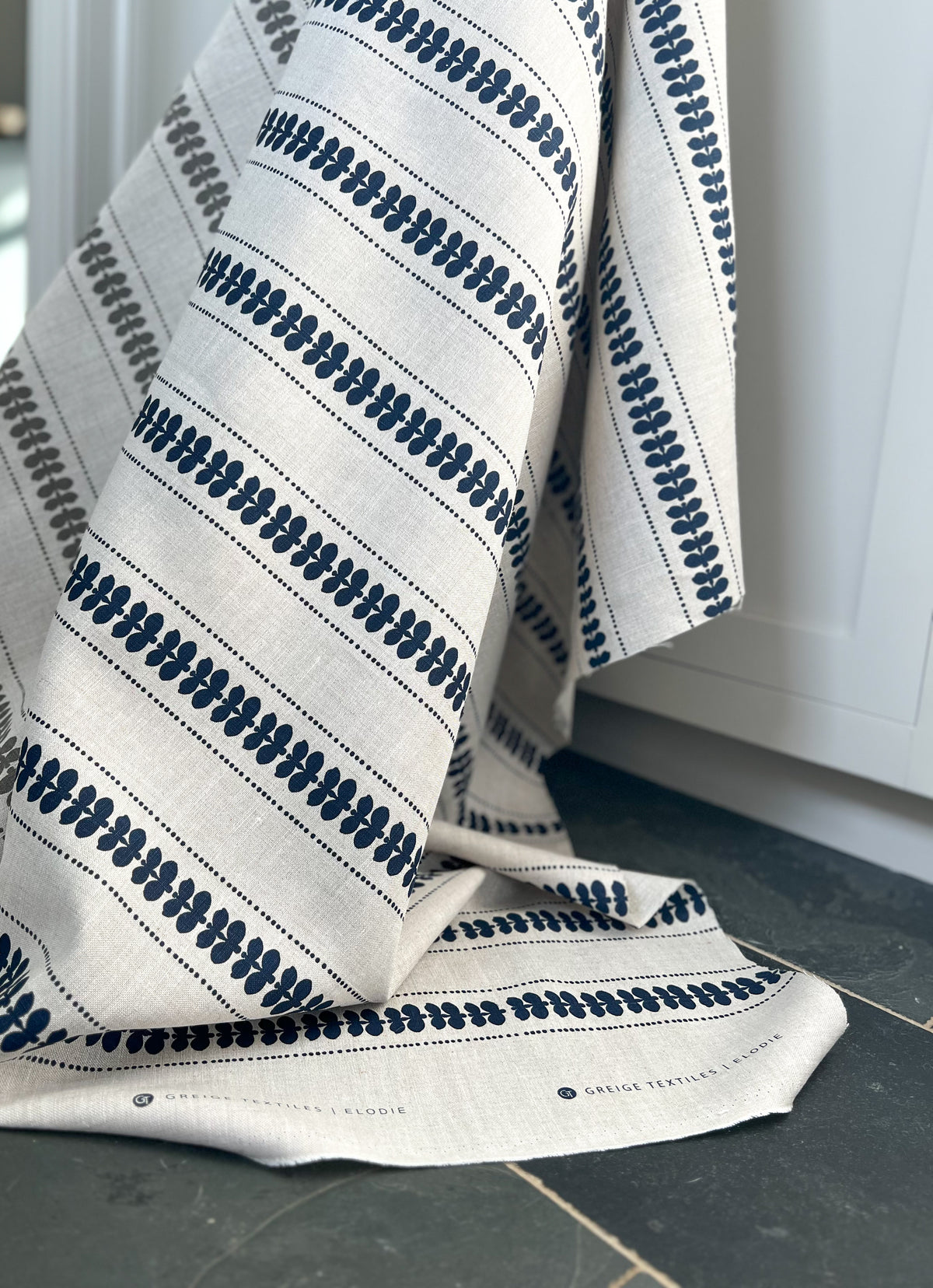 Artisan screen-printed linen with Elodie’s floral stripe design — delicate botanical elements arranged in a subtle, repeating rhythm. grey on oatmeal . Hand-printed by artisans, Greige Textiles&#39; fabrics and wallpapers add a refined touch to any interior.